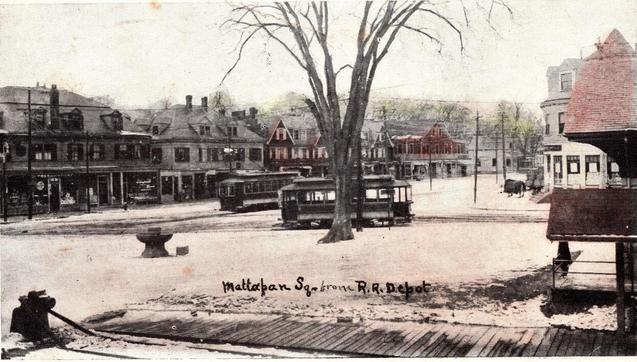 A snowy street scene at Mattapan Square shows early electric trolley cars stopped near the railroad depot, with storefronts and wooden buildings lining the square under bare winter trees.