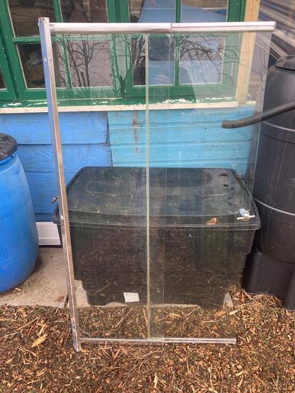 A shower screen is leant up against a rectangular water tank in front of a blue shed with green windows. The water tank holds rainwater harvested from the roof. The shower screen is approximately 140 centimetres high and 80 centimetres wide. The screen has two panels and the second panel has a metal handle and slips inside the first panel on runners. The screen will be fitted as an end window and horizontally so that the second screen will effectively become a drop-down window to ventilate the shed.
