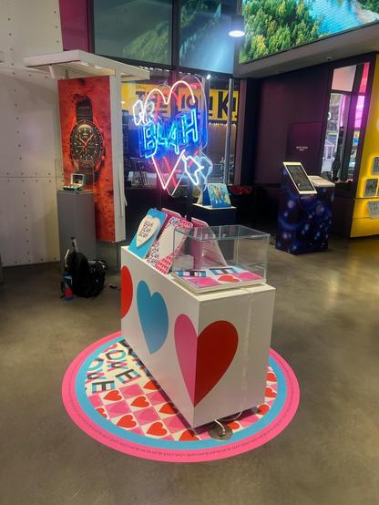 A retail display in a watch store with a neon sign over it with a heart and arrow through it. The sign has "BLAH" in blue through the heart.