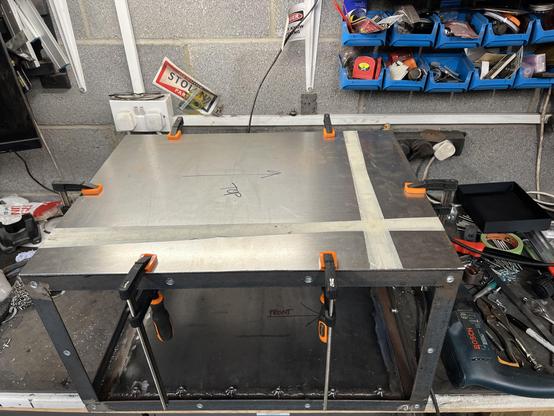 Metal frame sitting on a work bench. A sheet of metal cut to size sits on top and 6 clamps hold it in place.