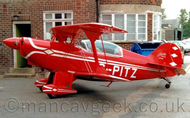 Side view of a small, red, single-engined bi-plane light aircraft parked facing to the left.
The plane has white stripes running along the body, with white registration "G-0PITZ" on the lower rear fuselage.
The red spats on the main wheels under the wing also have a pair of white stripes.
There are white "Pitts Special" titles on the tail, next to 3 white diamonds stacked vertically.
A large bubble canopy encases the 2 seat cockpit.
Grey concrete apron fills the foreground, with  a brown brick Art Deco building with white-framed windows stretches across most of the background from the left.
A green hangar with cars parked in front is visible on the right, under flat grey skies.