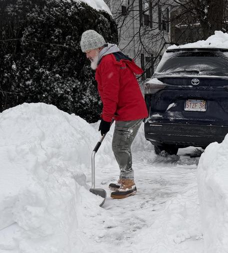 Old white man, white beard, in need of ibuprofen shoveling out waist high snow that has been packed in by snow plows.