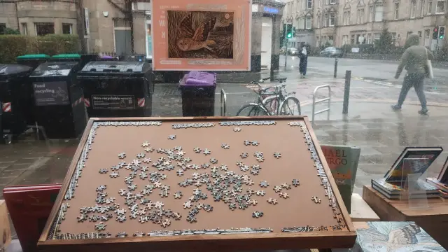 Angled puzzle table with a partially completed jigsaw on it, in the window of the Edinburgh Bookshop