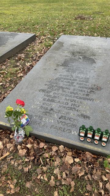 Das Foto zeigt einen großen, grauen, flachen Grabstein auf einem grasbewachsenen Friedhofsgelände mit herbstlichem Laub. Der Stein trägt eingravierte Inschriften auch FENJA PARLOW. Vor dem Stein liegen zwei gelbe und eine rote Rose mit einem Anhänger mit Regenbogenfahne sowie einem Broilersanhänger. Daneben stehen vier grüne Fläschchen Jägermeister nebeneinander. Links ist ein weiterer Grabstein sichtbar.