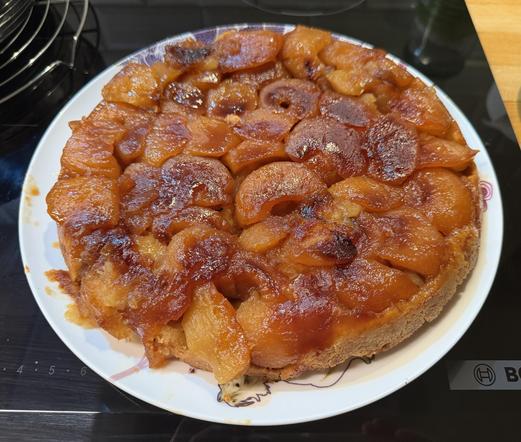 Une tarte tatin posée sur une assiette blanche qui est sur une plaque à induction. On voit bien les pommes encore bien dessinées et très caramélisées et très colorées. Elle donne vraiment très envie.