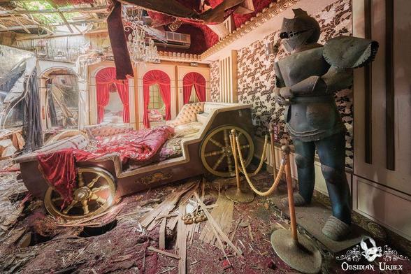 An abandoned themed suite shows a bed resembling a carriage, red drapes, and a knight statue. Debris is strewn across the floor and the ceiling is damaged.