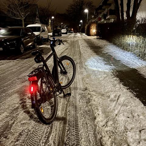Ein Fahrrad steht nachts auf einer verschneiten Straße, beleuchtet von Straßenlaternen. Autos sind an den Seiten geparkt, und Fußabdrücke und Reifenspuren sind im Schnee sichtbar.