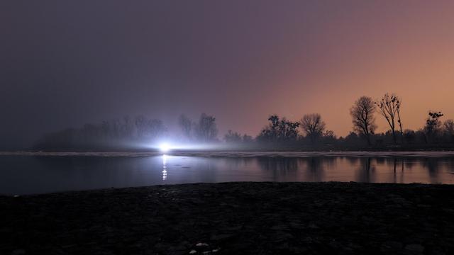 Hier sieht man von der Spitze des Rothehornpark auf die Elbe stromaufwärts. Sehr dominierend steht nahe dem gegenüberliegenden Ufer ein Auto, das dort mit eingeschaltetem Licht parkt und die ganze Szene überstrahlt.
Am unteren Bildrand sieht man die kopfsteinpflasterartige Befestigung des dortigen Ufers.
Große Teile der Elbe darüber sind noch nicht gefroren, allerdings durch die lange Belichtung nur als glasige Fläche zu erahnen.
Am gegenüberliegenden Ufer stehen Bäume und Büsche, die sich schwarz vor dem Himmel abheben, der wieder blassrosa bis orange leuchtet, wo die Stadt seitlich einstrahlt.