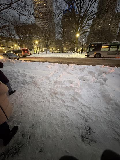 A snowy urban scene at dusk, featuring a snow-covered sidewalk and road. A couple of vehicles, including a bus and a car, are visible near buildings. Streetlights illuminate the area, and several trees are bare of leaves.