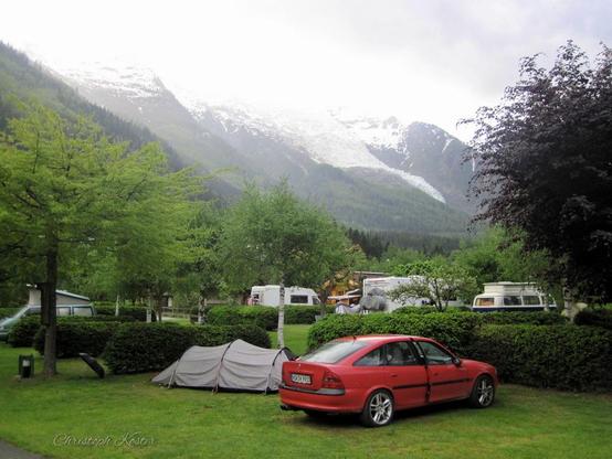 A vibrant red hatchback car is parked on a grassy campsite, surrounded by lush green trees and neatly trimmed hedges. In front of the car, there is a small, grey camping tent set up on the grass. The campsite is nestled in a scenic valley, with towering, snow-capped mountains visible in the background, partially shrouded in mist. The scene is tranquil, with several other campers, cars, and caravans scattered around the area, suggesting a peaceful outdoor getaway in a mountainous region. The overall atmosphere is serene and inviting, ideal for camping and nature enthusiasts.