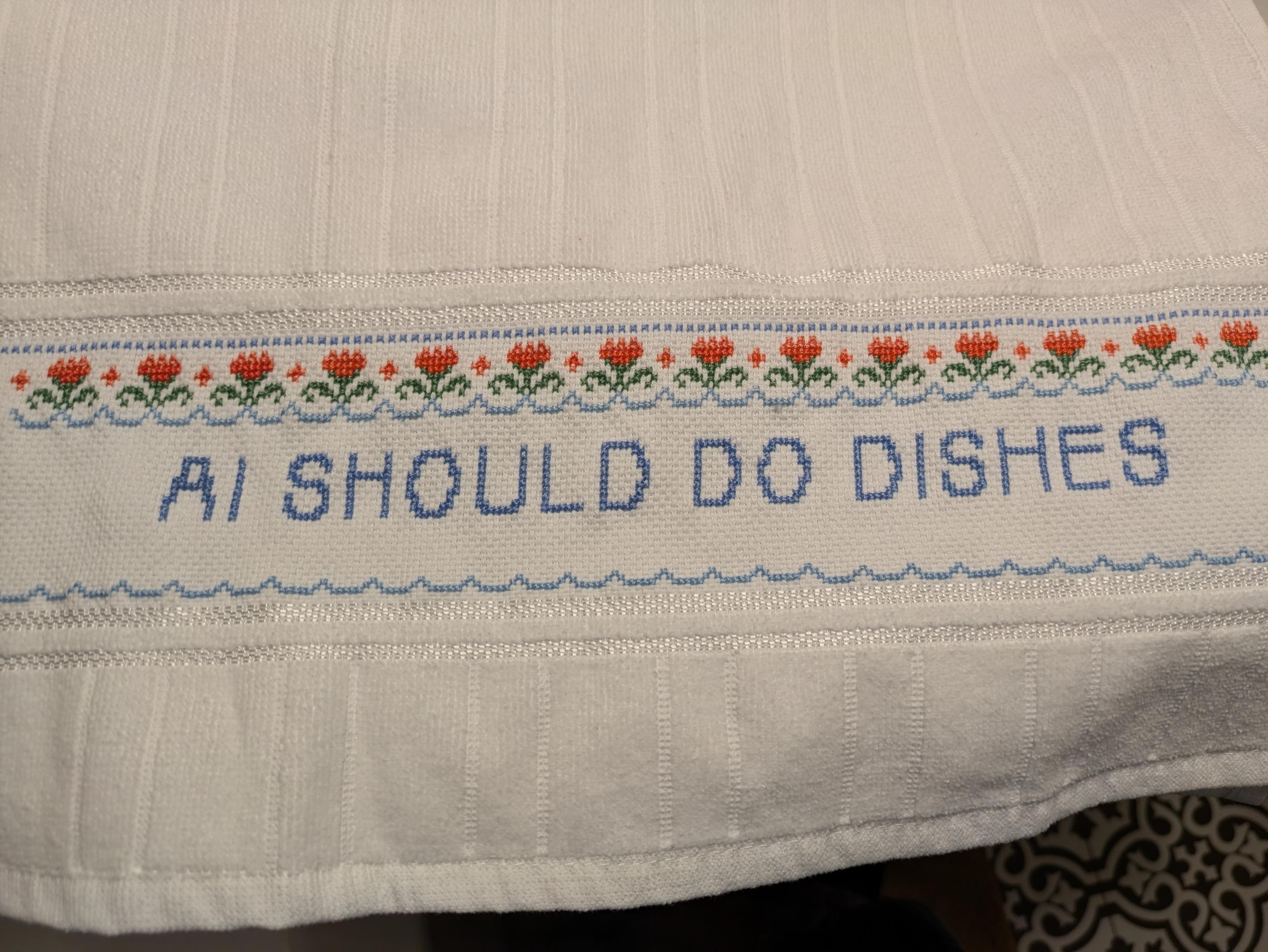 A white dishtowel with an embroidered panel. It features a row of coral flowers and some blue bunting. The message reads: "AI SHOULD DO DISHES."
