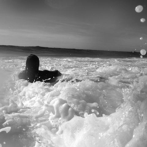 das quadratische schwarz-weiß-foto zeigt eine person im meer, von hinten gesehen. die person trägt einen dunklen anzug und kämpft sich mit ihrem surfbrett durch das schaumige wasser aufs offene meer. wellen sind im hintergrund zu sehen, und tropfen spritzen in der luft.