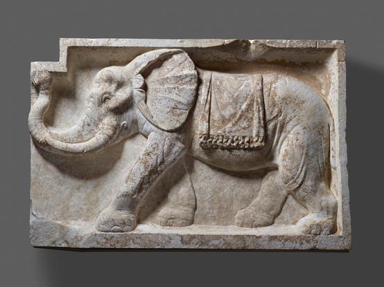 Description from The Getty: “Two rectangular architectural panels with plain moldings, each with the depiction of an elephant in relief. Both pieces are cut and chipped on their inner ends in such a way as to suggest that the elephants faced each other in an architectural setting. Small rectangular cut-away areas appear above the end of the trunk of each elephant. They stride across the panels with their trunks curled in a salute and with fringed ceremonial blankets on their backs. Incised crosshatching is used to convey the roughness of the hide. Unknown artist/maker (Roman); Pair of Architectural Reliefs with Elephants, A.D. 80–100; Italian marble; The J. Paul Getty Museum, Villa Collection, Malibu, California, 71.AA.463.”