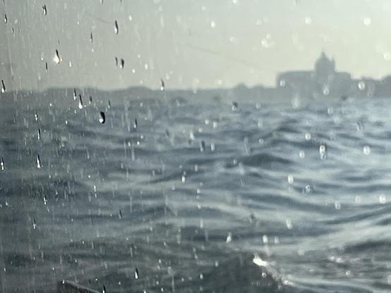 Colour photo taken through the window of a vaporetto water bus, looking out over a very wide canal of choppy waves, with the blurry shadowy skyline of an island, dominated by a big domed church under a milky grey sky, on the horizon line in the distance. The boat is quite low in the water, and the perspective makes it look like the waves are almost lapping against the window, which is dotted with sharp-focus water droplets that are catching the sunlight.