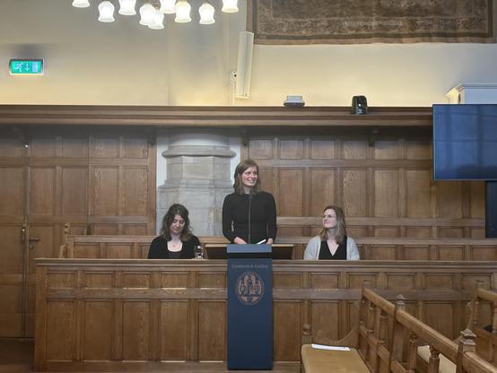 Aletta staat in het groot auditorium met mooie houten banken voor een logo van Universiteit Leiden, naast haar zittend haar paranimfen Liselotte en Nienke
