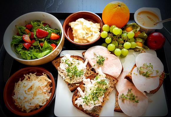Auf einem Tablett sind verschiedene Speisen vorbereitet. Ein kleine Schale Cole Slaw, Salat von Babyspinat und Erbbeeren, ein Klacks Kartoffelsalat, Orange, Trauben, Physalis, Pflaume, ein Pudding und ein Brettchen mit belegten Broten, die mit Kresse ganiert sind.