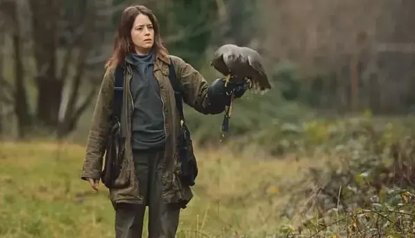 A still from the film H Is For Hawk. A white woman walks through the countryside holding a goshawk on her left wrist.