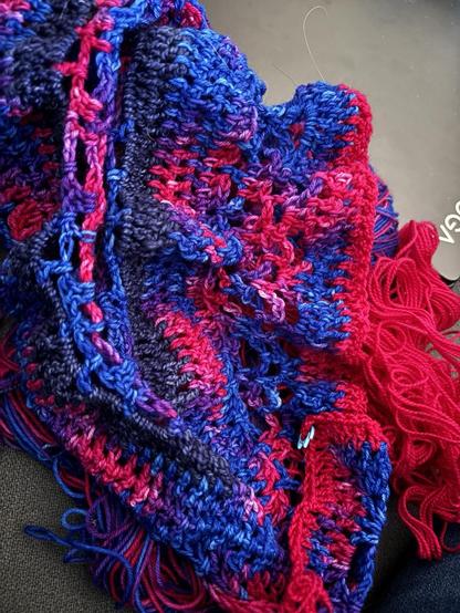 A picture of a crocheted shawl in progress. The colors are a saturated bi pride colorway. It is on a gray couch.