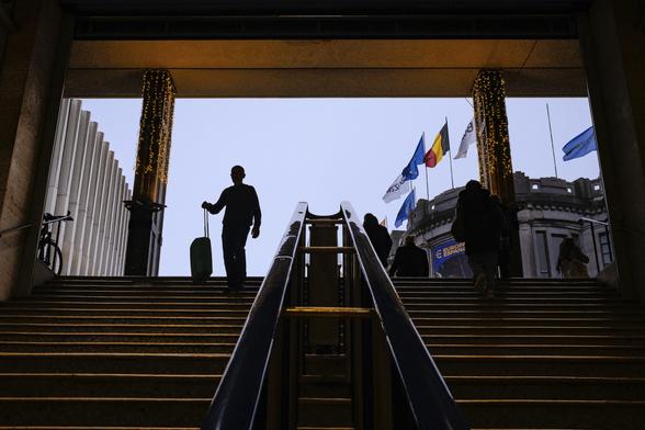 Look up shot of a man holding a suitcase, his silhouette contrasting on a light blue-grey sky, starting to descend stairs into a dark passage-way leading to a station. The image also shows a subtle play of colours responding to dominating shadows with touches of blues (sky, flags in the background) and some warm orange lines on the structure framing the stairs, on the edges of the steps and in a Belgian flag.