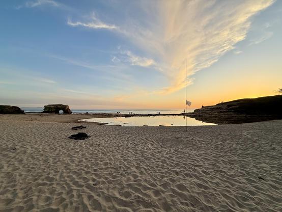 Photo from beach of Natural Bridges SB facing the pacific. The natural bridge is on the left, cliffs on the right and the antenna set up in the middle on the beach.
The sun is almost setting, giving the blue, slightly cloudy sky a nice orange touch at the horizon.