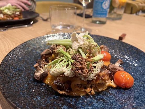 Ein lebendiges Gericht, das auf einem dunkelblauen Teller präsentiert wird, mit Nudeln, die mit Sauce überzogen sind und mit verschiedenen Zutaten wie gerösteten Kirschtomaten, knusprigem Gemüse und Pilzen belegt sind. Der Hintergrund umfasst ein Glas Wasser und verschwommene Speiseelemente.