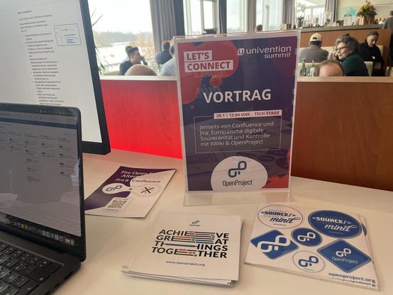 OpenProject booth at Univention Summit 2026 with laptop, flyers, stickers, and a sign promoting a joint talk with XWiki.