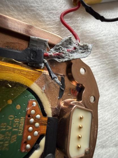 Close-up image of the internal components of an electronic device, showing a circuit board with visible wiring, damaged insulation, and solder points. The area includes exposed wires, a connector with gold pins, and a copper casing.