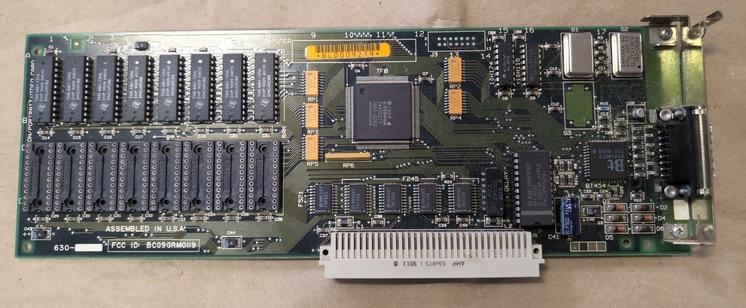 A long NuBus card lying on a piece of cardboard. It has a single external D-sub connector. On the left are eight chips soldered to the board with eight empty chip sockets below them. ASSEMBLED IN USA is legible above the FCC ID: BCG9GRM0119.