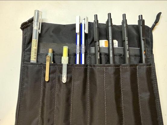 A foldable pencil case, laid open on the table, with eight slots for pens, four on each side. All the pencils on the right have been partially withdrawn. On the right are three  Rotring Rapid Pro (one pen, a 0.7mm and a 0.5mm pencil) and a Uni Kura Toga 0.5mm on the right. The pockets on the left side show a Muji 0.5mm ballpoint pen and two Tombow Mono Zero erasers (round and square). Still fully seated on the left are a Muji highlighter and a Tactile Turn Nautilus.