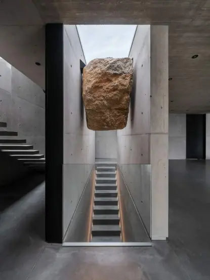Photo of the inside of a very minimalistic house. In the center of the floor are stairs leading downward with walls on the sides. Sandwiched between the walls, and thus right above the stairs, is a boulder.