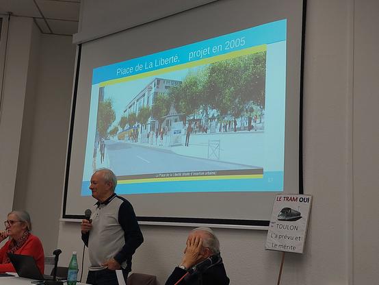 un vieux homme blanc en train de montrer un diapo sur lequel figure une visualisation du projet tramway de Toulon de 2005. il y a moins de voitures et plis d'arbres et généralement la place liberté est visiblement plus calme qu'elle ne l'est aujourd'hui