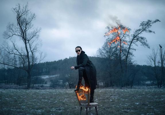 A figure in dark clothing sits on a wooden chair engulfed in flames, with a smoky, dramatic landscape in the background.