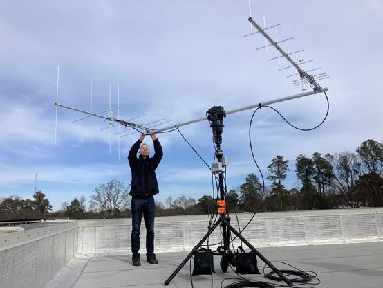 A man adjusts an antenna on a large remote controlled array. He’s standing on the roof with the sky behind him.