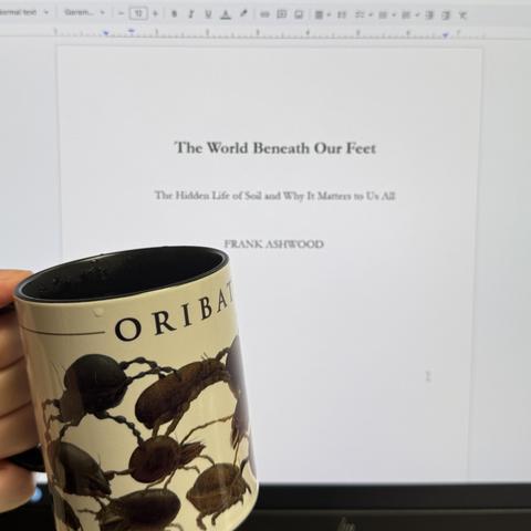 A photo of a computer monitor with the title page of a book, entitled 'the world beneath our feet' by Frank Ashwood. In front of the monitor is my hand, holding a mug with images of many hard-bodied oribatid mites on it.