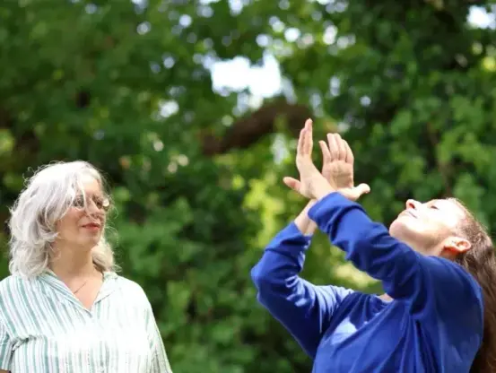 Photo de deux femmes, l'une regardant l'autre en train de signer, visage vers le ciel, yeux clos, en pleine nature