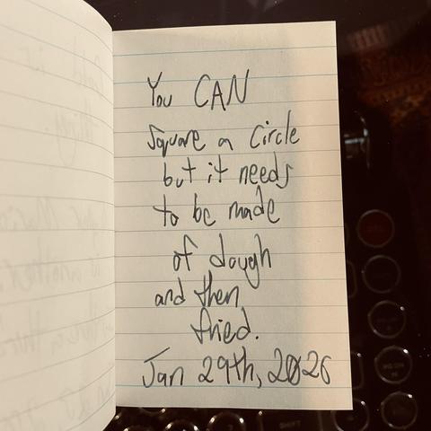 Close up shot of a tiny ruled notebook on a glasstop desk. Someone's written something on the page, "You CAN square a circle but it needs to be made of dough and then fried." The note is dated, Jan 29th, 2026.