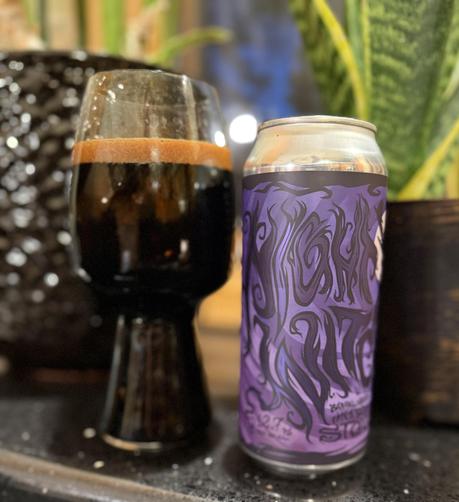 A stout glass full of pitch black dark beer. A the silver can is wrapped in a purple label with wiggly black lettering spelling out Night Witch 2025