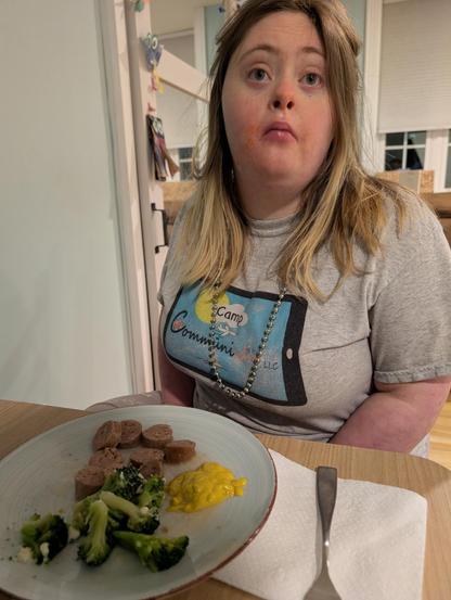 A young woman with Down syndrome sitting at a table with a serving of sausage (cut up), broccoli florets and a dollop of mustard.