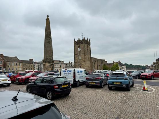 A cobbled market square is filled with parked cars of various colours, with a white service van marked “Job Done” prominently visible. A tall stone obelisk with a round finial rises from the centre of the square, while a historic stone building with a clock tower and crenellated roof stands behind it. The surrounding area features rows of traditional stone and brick buildings with sash windows, typical of a small historic market town. The sky is overcast, casting a grey tone across the scene, and a single traffic cone marks a reserved parking space near the foreground. Trees and low hills are visible in the distant background.