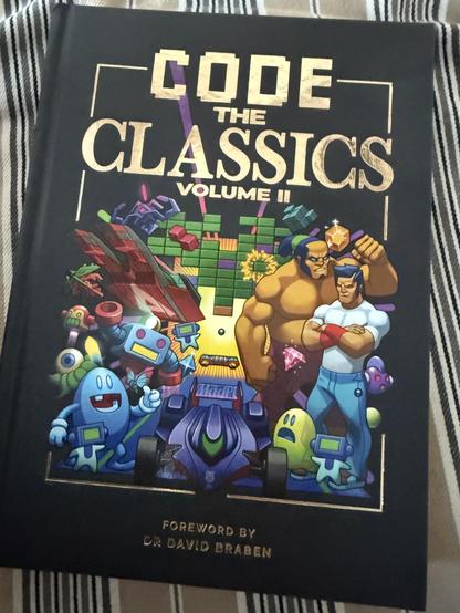 Photo of the cover of a book. The book has a dark border, and across the top is the title in gold: Code the Classics volume II. 
In the middle of the book are multiple cartoon drawings of characters from the games included in the book.