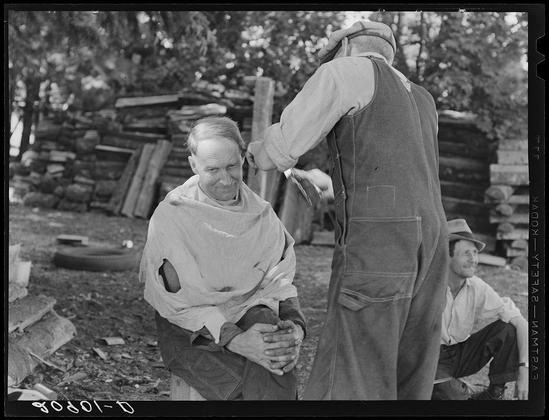 The image is a black and white photograph depicting three individuals in what appears to be an outdoor setting with natural foliage in the background. The central figure, presumably male based on physical characteristics visible, sits down wearing ragged clothing that includes torn sleeves and patches over holes or tears. His posture suggests discomfort as he gazes downward with his head slightly bowed.

Behind him stands another person who is actively engaged in cutting hair; this individual wears a cap and dark-toned workoveralls typical of manual labor attire from the early to mid-20th century, indicative of rural working conditions. The standing figure's attention seems focused on their task while interacting with the seated man.

To the right side of the frame is a third person who appears relaxed in posture; this individual sits down and wears casual clothing consisting of jeans and a shirt or sweater. This person watches the interaction between the other two figures, adding to the social context within which these events are occurring.

The environment suggests an informal outdoor setting with stacked wood behind them and scattered debris around their immediate area. The photograph carries historical significance as it is captioned "Bean pickers barbering each other," hinting at a moment of camaraderie or necessity among laborers during harvest time in the Pacific  [...]