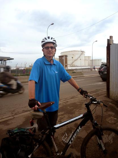 A person standing beside a black Trek mountain bike on a city street, wearing a blue polo shirt, black gloves, and a white bicycle helmet. In the background, large white oil storage tanks and a road with passing vehicles are visible under a cloudy sky.