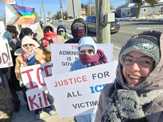A small group selfie of 8 men and women holding handmade protest signs as they stand on the sidewalk next to a busy road.  They are bundled up due to frigid temperatures.  Of the signs that are visible, one says,"JUSTICE FOR ALL THE ICE VICTIMS."  Another reads, "TRUMP'S ADMINISTRATION IS NOT GOVERNMENT.  IT IS A CRIMINAL ENTERPRISE."  Another says, "ICE KILLS." Another reads, "ICE OUT FOR GOOD."