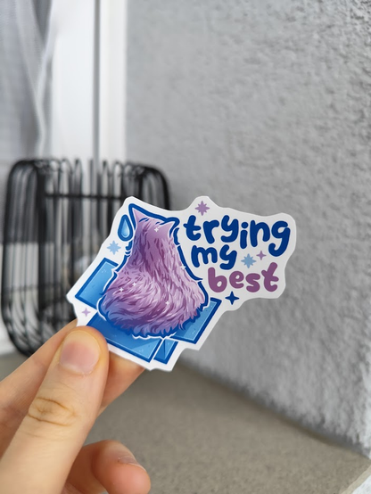 hand holding up a sticker of a fat kitty sitting in a box that's too small, titled "trying my best"