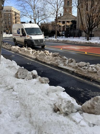 A white USPS delivery van with the logo and the words “United States Postal Service” and “We Deliver For You.” is driving on a wet, cleared road bordered by plowed snowbanks. Large chunks of dirty snow line the sidewalk and separate the bike lane from the general travel lane, while the street features a red-painted bus lane with yellow markings. In the background, leafless trees, a brick building, and a church with a tall clock tower are visible under a bright blue sky.