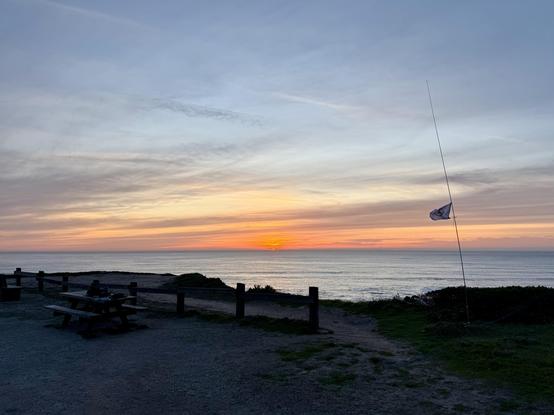 Sunset over the Pacifica on the left is a bench with some radio gear, on the right is a vertical antenna with a POTA flag flying in the wind to the left.