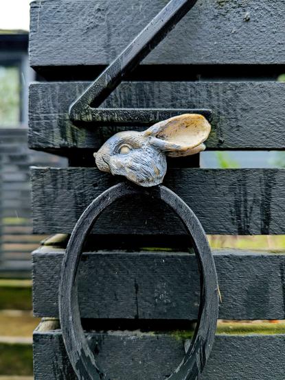 A photo of the weathered plastic head of a toy rabbit wedged between black metal numbers mounted on the painted black wooden slats of a fence, with blurred greenery and a house partially visible in the background.