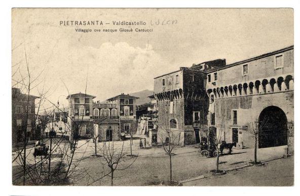 This black-and-white historical photograph depicts a quaint village scene, labeled as "Pietrasanta - Valdicastro" with the notation that it is where the author Gioseù Carducci was born. The image shows European-style buildings, including one with decorative arcades on its upper floors and another featuring an arched entranceway.

A horse-drawn carriage is seen parked by the rightmost building, adding to the historical ambiance of the scene. Bare trees in the foreground suggest that this photo may have been taken during late autumn or winter. The overall setting appears peaceful, with no visible people present on the street at the time captured in the image.