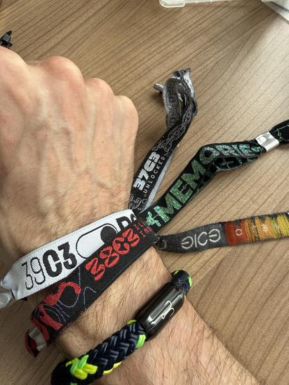 Das Bild zeigt eine Hand mit mehreren bunten Armbändern, darunter Stoff und gewebte Stile, die vor einer Holzoberfläche ausgestellt sind. Die Armbänder verfügen über verschiedene Designs und Texte, alle vom Chaos Communication Congress.