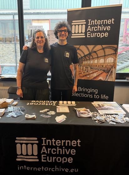 A man and a woman stand in front of the Internet Archive Europe stand at FOSDEM in Brussels